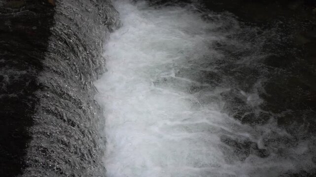 Close-up of a fast-flowing mountain river cascading over a natural weir. Whitewater foams intensely as it splashes down the rocky ledge. Bright daylight, dynamic and powerful nature scene.