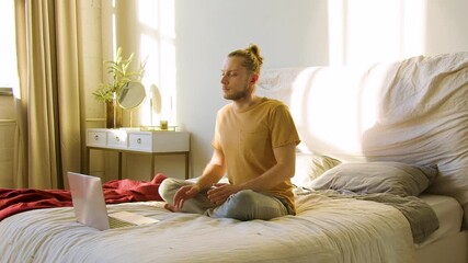 Dolly shot of a young man chooses an online meditation tutorial, sits in a lotus position and begins to meditate. He finds Pilates exercise videos on the internet to get ready to meditating practice.