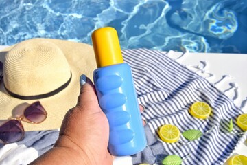 Sunscreen in hand and straw hat on beach towel by the pool. Protection for Summer vacation in the...