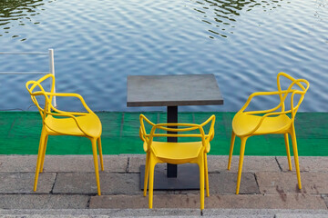 Table and yellow plastic chairs on the shore near the water. Empty cafe area on the embankment