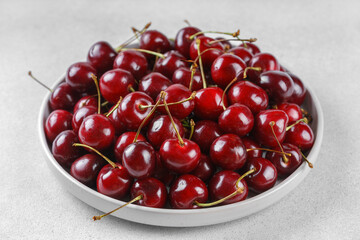 Heap of fresh ripe red sweet cherry with stems in white plate on light textured background. Natural...