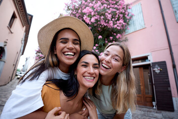 Three cheerful female friends laughing and embracing each other while enjoying their vacation on a picturesque street adorned with vibrant pink flowers, capturing moments of joy and togetherness
