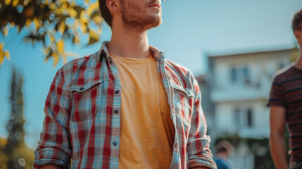 A young man with a beard wearing a plaid shirt and yellow t-shirt is gazing upward in an outdoor setting.