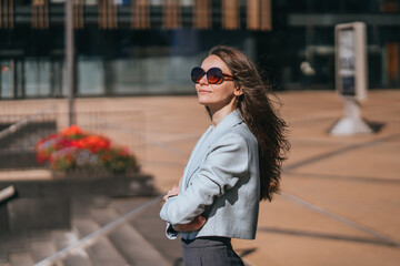 A woman in sunglasses embraces a laptop as she stands in a sunlit plaza surrounded by towering glass buildings.