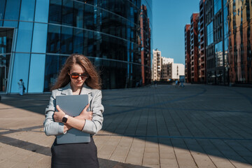A confident woman in a green suit sits on modern urban furniture with a laptop, embracing the day with a bright smile.