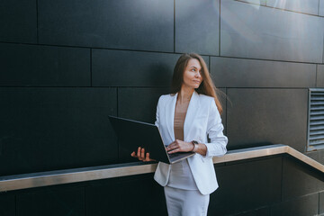 A woman in a white suit stands confidently with a laptop against a dark modern wall.