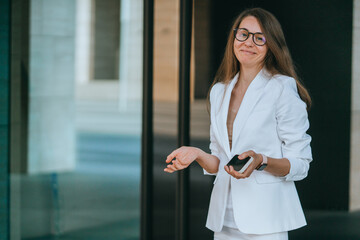 A woman in glasses and a white suit gestures expressively outdoors.