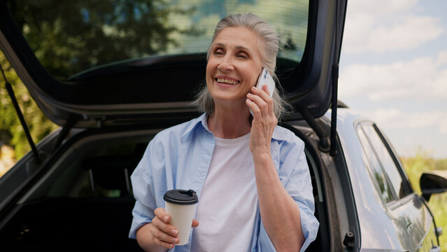 Happy smiling Caucasian retired business woman female senior mature automobile car driver businesswoman freelancer talking mobile phone speaking smartphone drinking coffee cup traveling outside nature - Powered by Adobe
