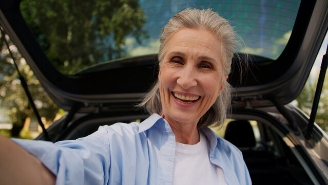 Happy cheerful aged Caucasian grandmother woman female senior car auto driver mature lady vlogger blogger influencer recording vlog blog travelling talking webcam greeting waving hand outdoors nature