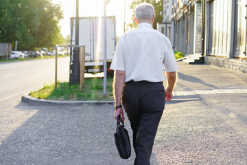 Elderly man walks along city street carrying a briefcase during late afternoon light in urban...