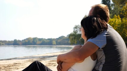 Happy pair hugs during sitting on wooden log on beach. Romantic couple admiring sea landscape at sunny day. Lovers spending time together at resort. Concept of summer vacation or holiday. Slow mo