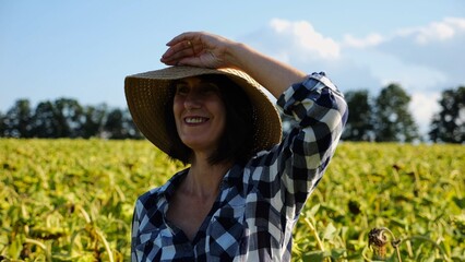 Happy smiling female farmer looks into camera standing near sunflower field. Portrait of adult beautiful agronomist in straw hat with yellow plantation at background. Agricultural business concept