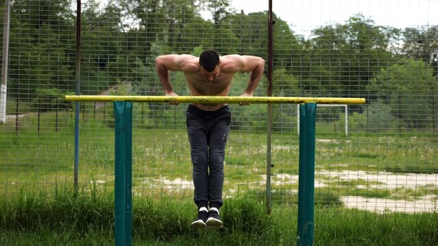 Hardy shirtless sportsman doing exercises on parallel bars at nature. Strong and muscular man working out at sports ground. Young athletic guy exercising outdoor. Concept of active healthy lifestyle