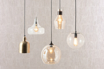 Assorted hanging pendant lights in sculptural clear glass and brushed metal finishes with exposed filament bulbs, set against a textured beige wall, evoking a modern eclectic style with warm ambiance