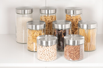 A close-up shot of a variety of seasonings and cereals stored in Clear Glass Jars Glass jars with stainless steel lids neatly arranged on white shelf, filled storage containers on a white wooden shelf
