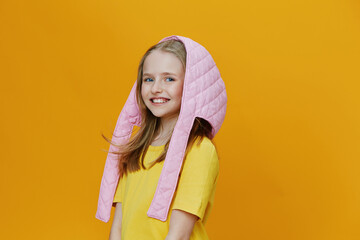 Happy girl wearing a pink quilted hood and yellow shirt against a vibrant yellow background,...