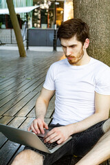 Young man in white t-shirt and shorts sitting on wooden floor, leaning against a tree, working on laptop. Frontal view, relaxed posture, natural outdoor setting.
