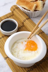 Half-boiled egg in bowl, soy sauce, chopsticks and toasted bread on white marble table, closeup. Traditional asian breakfast
