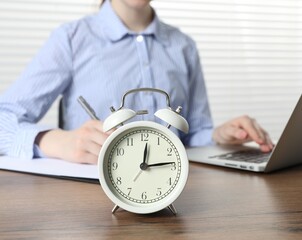Deadline. Woman with laptop and clipboard working at wooden table in office, focus on alarm clock
