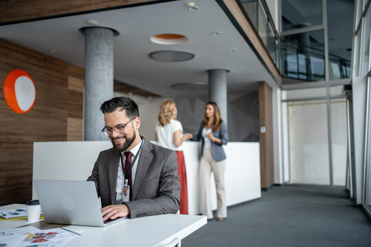 Businessman working on laptop in modern office lobby with coworkers chatting in background