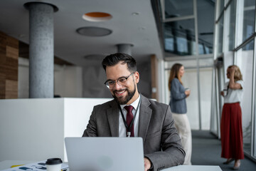 Businessman working on laptop in modern office with colleagues chatting in background