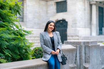 Smiling young Latina business woman in gray blazer and blue pants sitting relaxed near old stone...