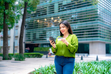 Young Latina woman in green blouse holding credit card and smartphone, smiling outside corporate...