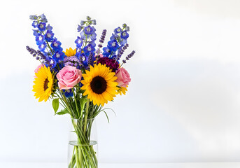 A vibrant bouquet featuring sunflowers, pink roses, and blue delphiniums arranged in a clear glass vase against a white background.