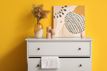 Vase with dried plant and painting on white chest of drawers near yellow wall