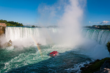 Scenic view of Horseshoe Falls at Niagara with a tour boat approaching through mist and a visible rainbow. A classic Canadian natural wonder on a sunny day.

