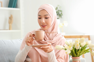 Beautiful young Asian woman in hijab with cup of tea sitting at living room