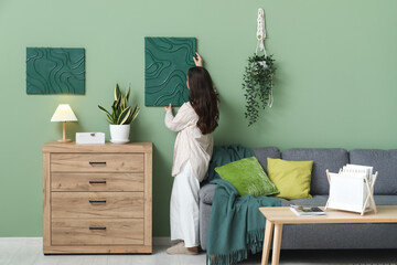 Young pretty woman hanging picture on green wall near chest of drawers in living room