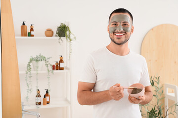 Handsome young happy man with cosmetic clay mask on face in bathroom at home