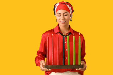 Young African-American woman with burning candles on yellow background. Kwanzaa celebration