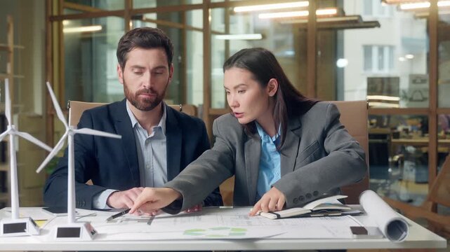 Japanese businesswoman inspector Caucasian businessman speak wind turbine model eco implementation renewable project document blueprint electricity engineering indoors office multinational meeting