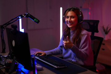 Asian young adult woman streamer content creator gamer live streaming from home studio giving thumbs up using computer headphones technology setup
