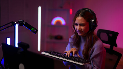 Young Asian adult streamer gamer plays popular online game using computer setup keyboard headphones microphone vibrant neon room
