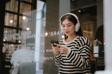 Fototapeta premium A cheerful young woman with braces wears headphones and smiles while video calling on her smartphone, enjoying a casual moment in an urban setting.
