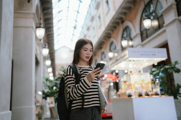 A modern young woman in a striped shirt stands in an urban mall, holding a laptop and smartphone,...