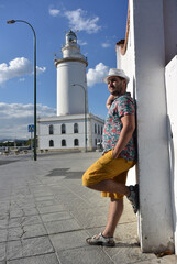Vertical street city travel photo. Mature male with beard, white hat, yellow shorts, floral print beach shirt stands near wall and Lighthouse La Farola. Concept of tourism, trip, summer vacation