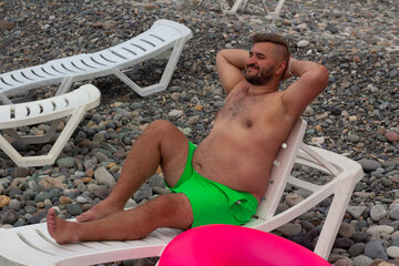 Horizontal travel photo. Full body mature tanned fat white naked man with beard, big belly, green shorts, pink swim ring sits and rests on plastic sun lounger on pebble sea coast. Tourism, relax, calm