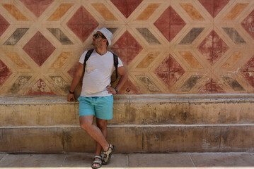 Street city photo. Mature man with stubble, sunglasses, blue shorts, white T-shirt, hat, sandals, backpack stands near old vintage wall of house with geometric motley shabby pattern. Trip, travel