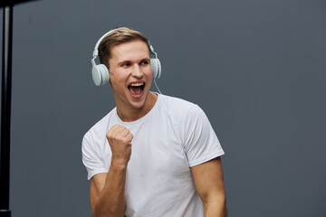 Excited young man wearing white t-shirt and headphones celebrating with clenched fist isolated on...