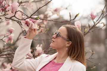 Magnolia woman spring: Lady in shades admires pink magnolia flowers on bright spring day outdoors.