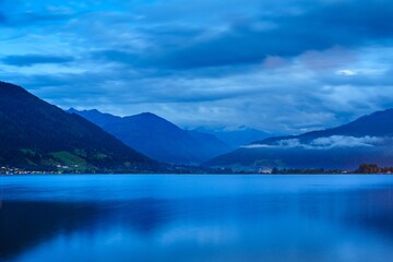 A tranquil dusk landscape featuring a calm lake that reflects towering mountains beneath a cloudy blue sky. Perfect for travel brochures and nature-themed publications.