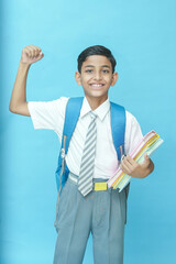Happy Indian schoolboy student holding books show strong arm gesture isolated on blue background,...