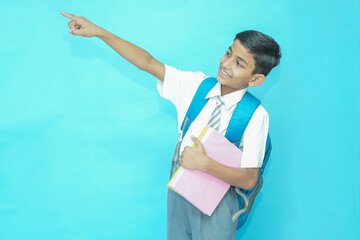 Indian student schoolboy wearing uniform holding notebooks and bag pointing at blank space isolated...