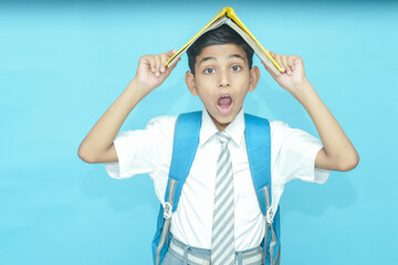 Surprised Indian primary school boy student with notebook on his head isolated over blue...