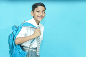 Happy Indian schoolboy wearing uniform and bag standing isolated over blue background, Education...
