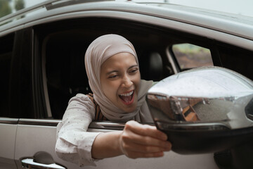 A happy woman adjusts the side mirror of her car while smiling gleefully, representing joy, human...